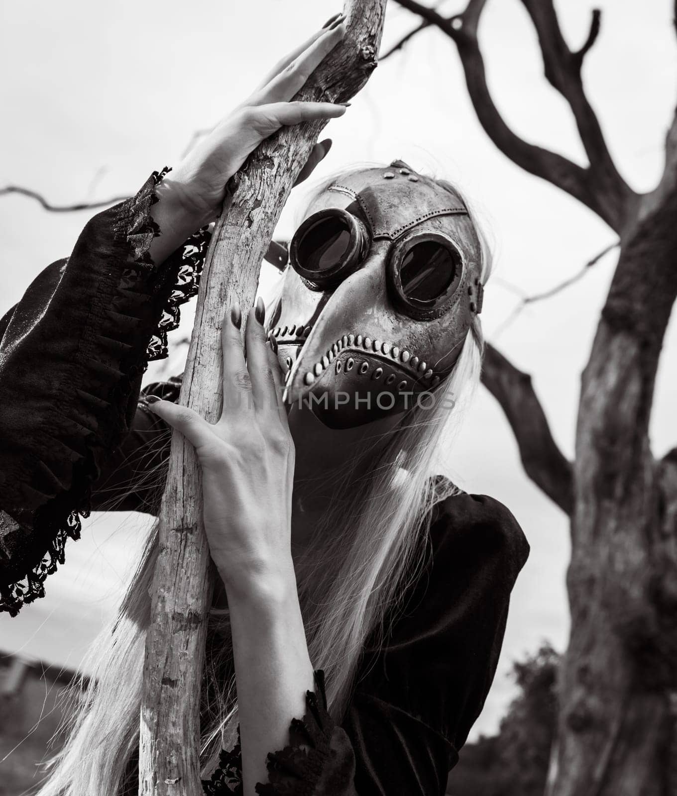Blonde woman in a crow mask posing near a withered tree in nature, creating an aura of mystery and beauty.
