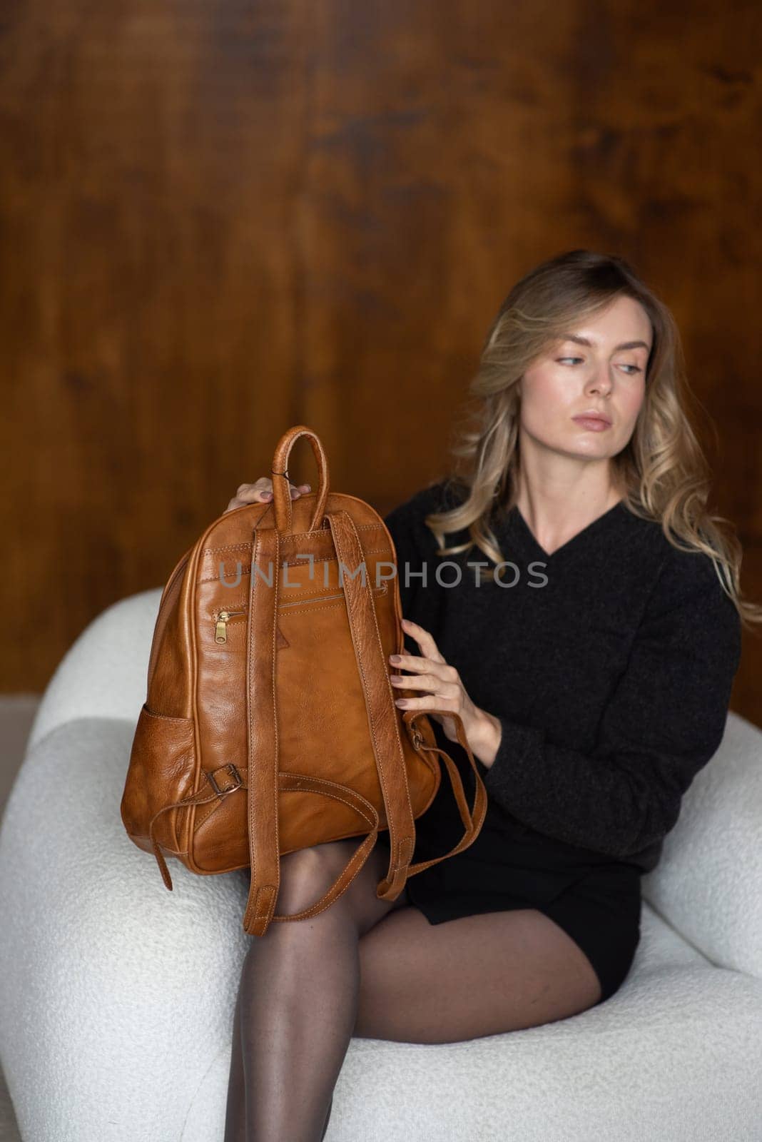 A girl in a warm sweater, a black short skirt, nylon tights, black loafers and gray socks is posing with a brown backpack
