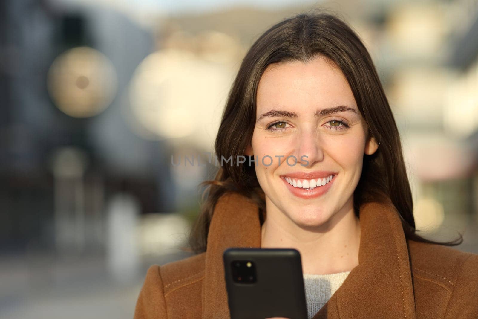 Front view portrait of a happy woman in winter holding phone looking at camera