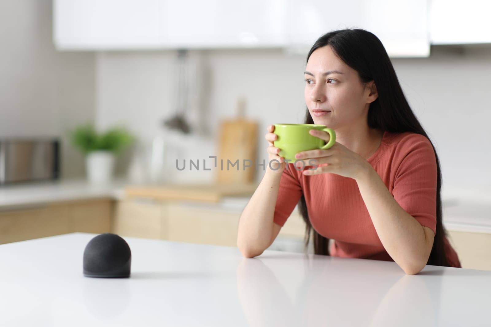 Relaxed asian woman drinking in a kitchen beside a smart speaker at home
