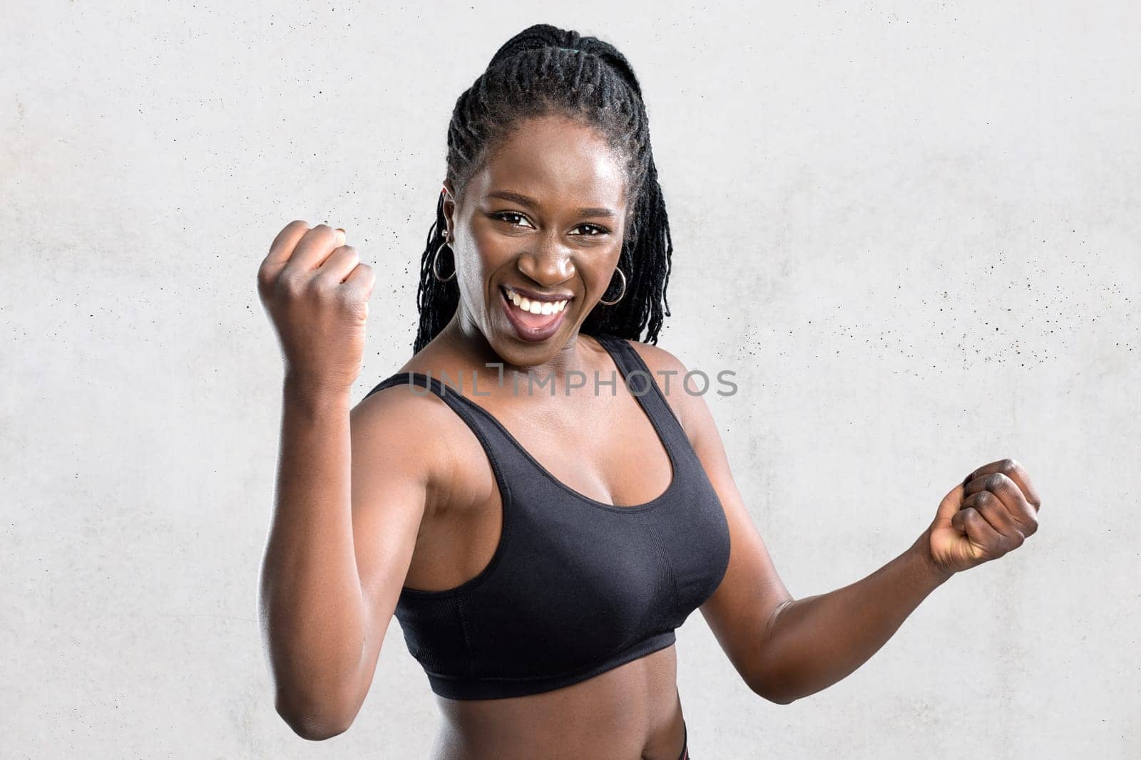 Close up portrait of sportive afro american teen athlete showing winning attitude.Young woman in sportswear raising hands against grey concrete background.