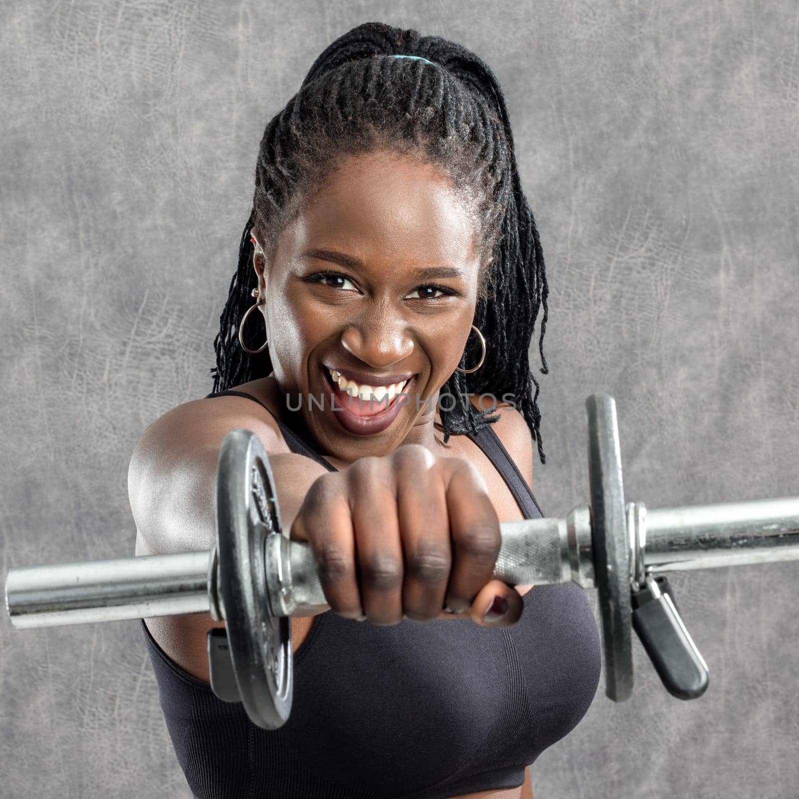 Close up portrait of sportive black teen with braids holding dumbbell in front of face. Young woman in sportswear with vigorous face expression against grey background.