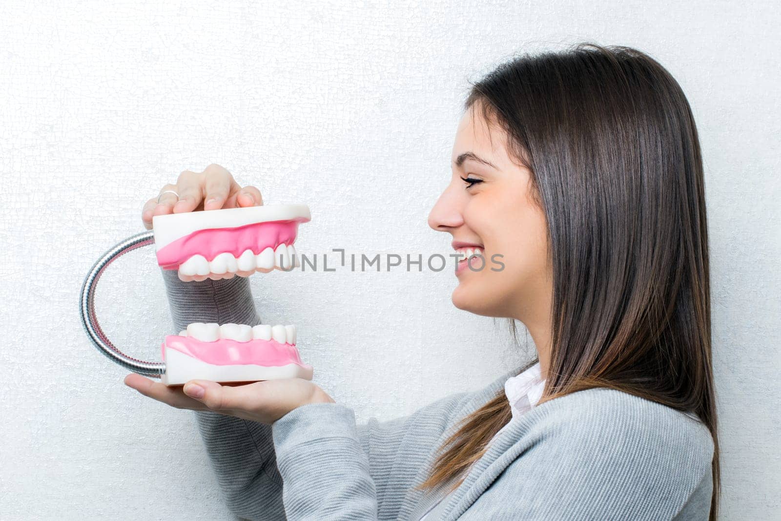 Close up portrait of attractive young girl holding oversize human teeth prosthesis.Side view of woman against light textured background.