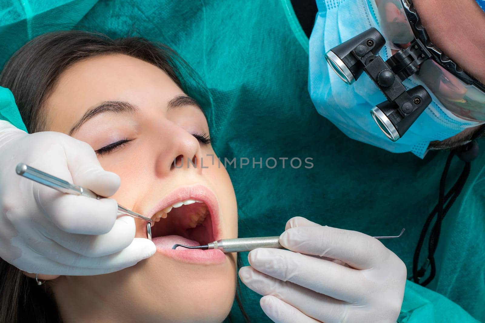 Close up of dentist wearing medical magnifying glasses working on female patient.Surgeon in green gown with mouth mirror and scaler.
