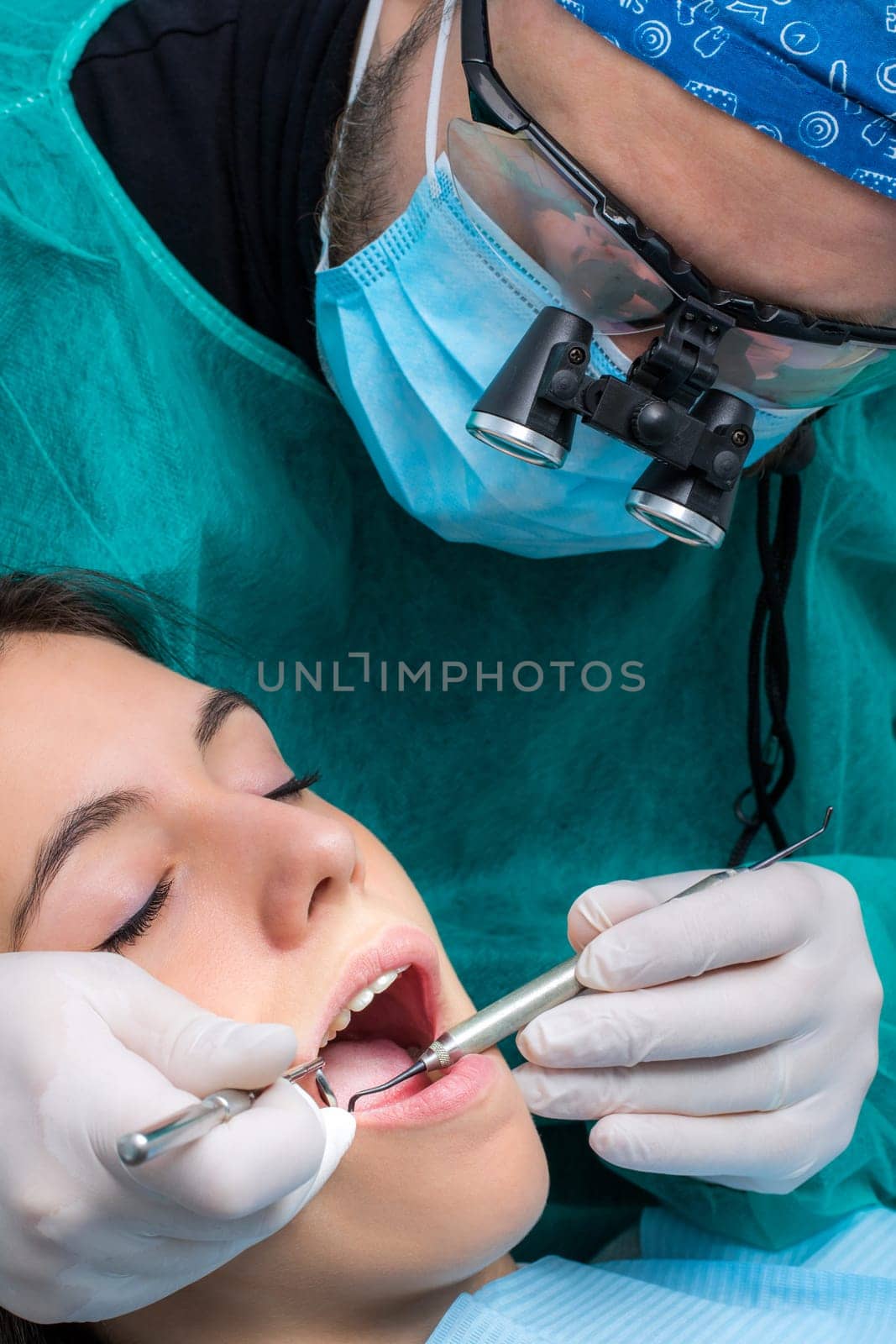 Close up of dentist wearing medical magnifying glasses working on female patient.Surgeon in green gown with mouth mirror and scaler.