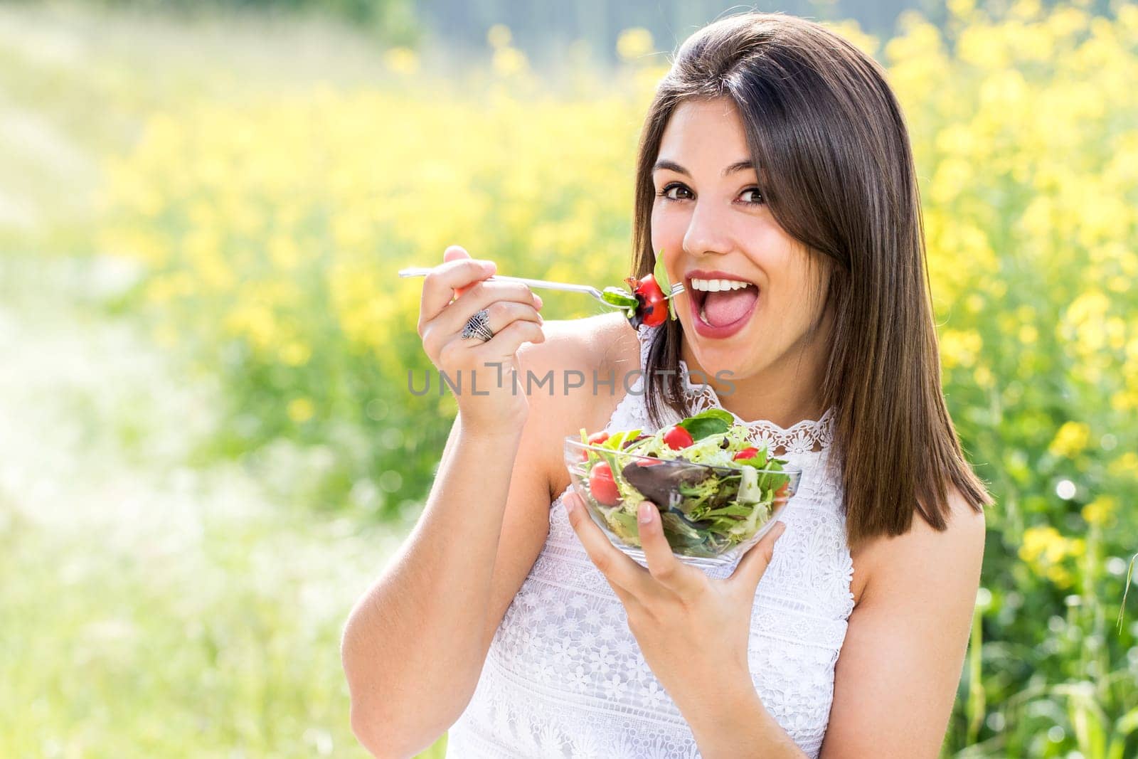 Close up portrait of attractive healthy young woman eating green salad outdoors.Girl holding salad bowl against yellow flower field.