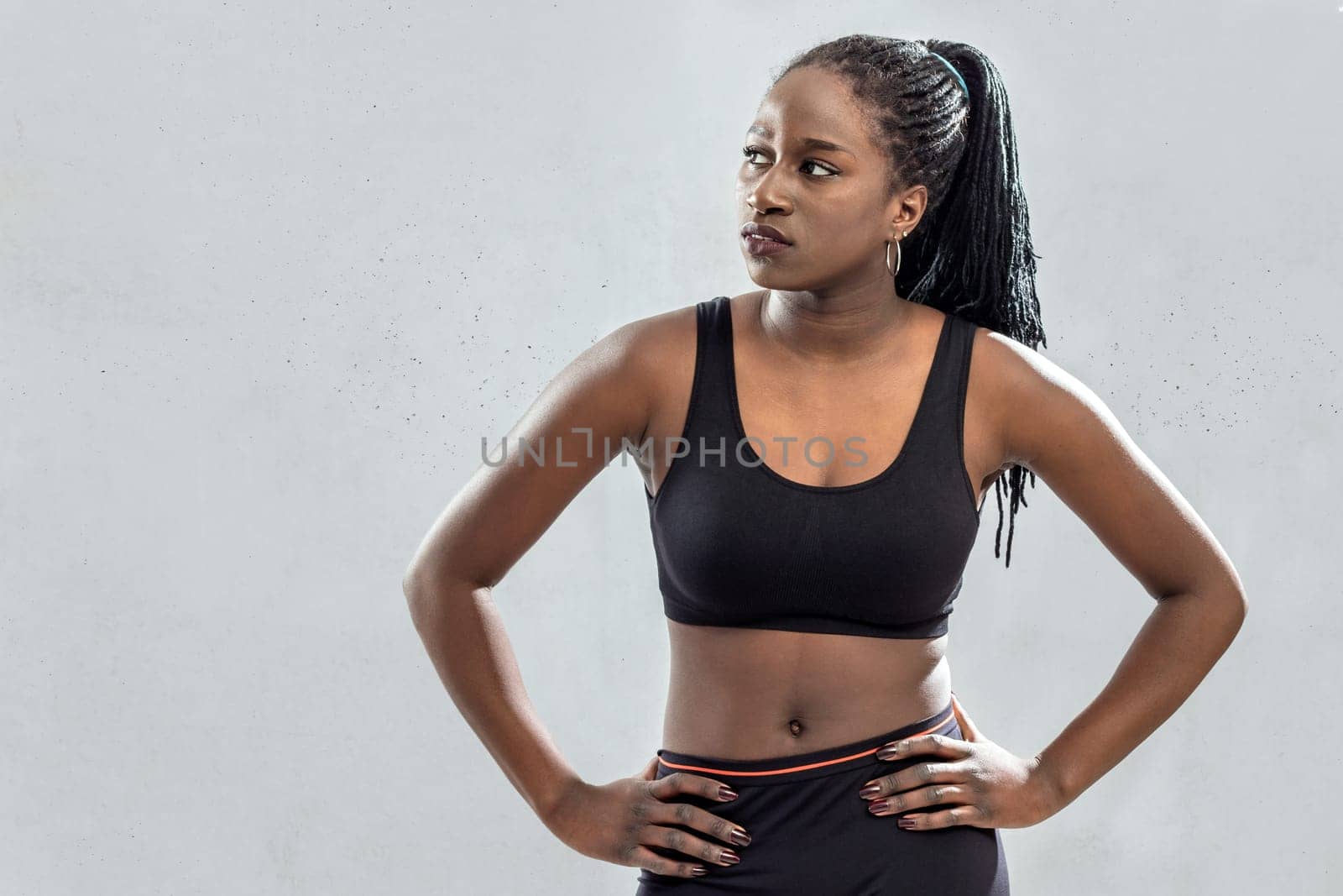 Close up portrait of muscular african teen athlete standing with hands on hips.Young woman looking aside against light grey concrete background.