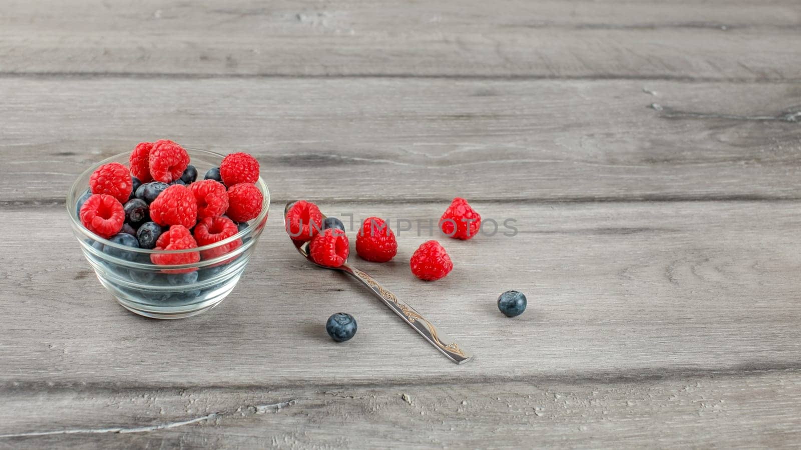 Glass bowl with blueberries and raspberries on top, with spoon and some spilled berries on the side placed on gray wood desk.