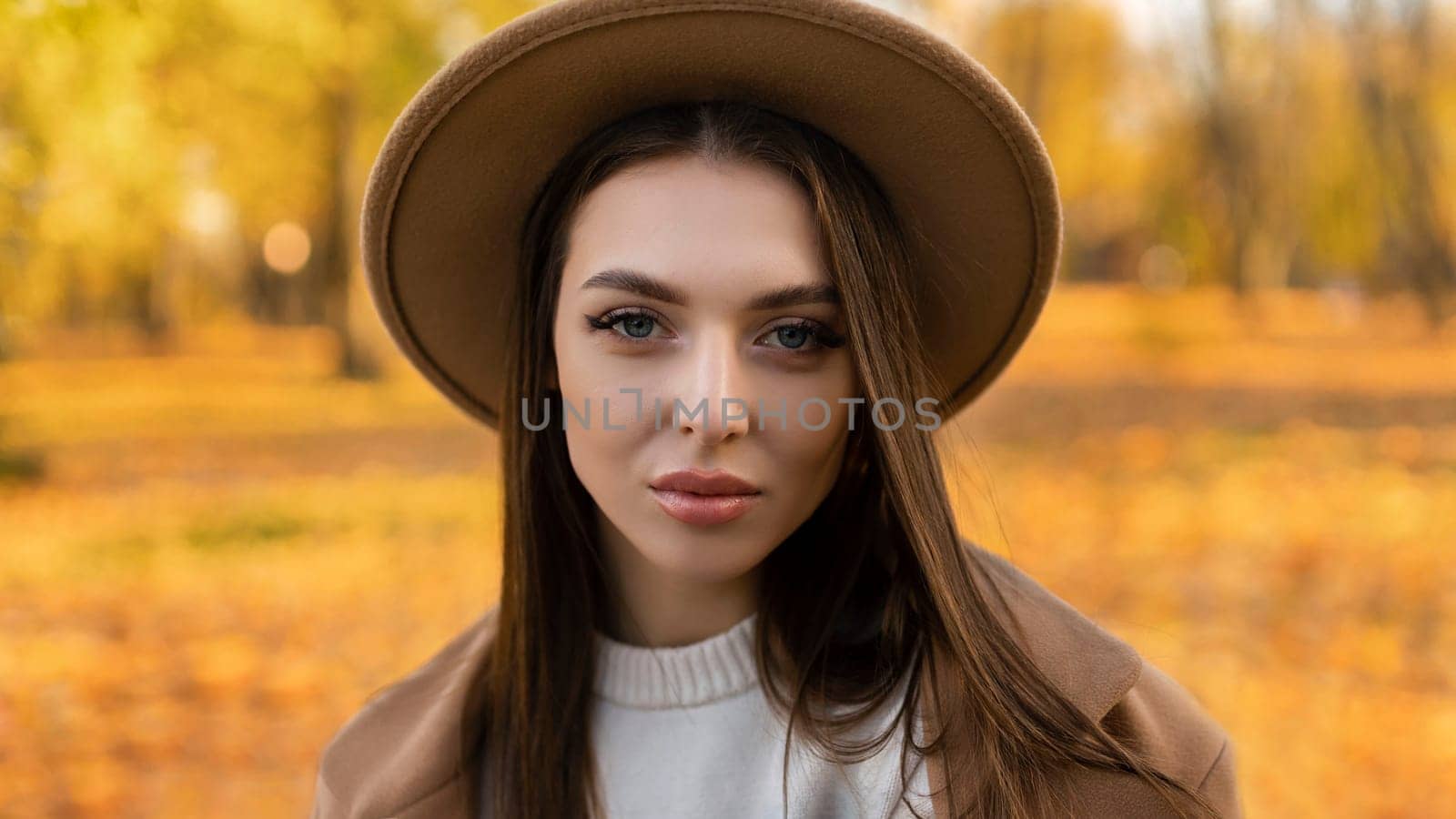 portrait of attractive stylish smiling woman with long hair walking in the park, dressed in warm brown coat autumn trendy fashion, street style wearing hat.