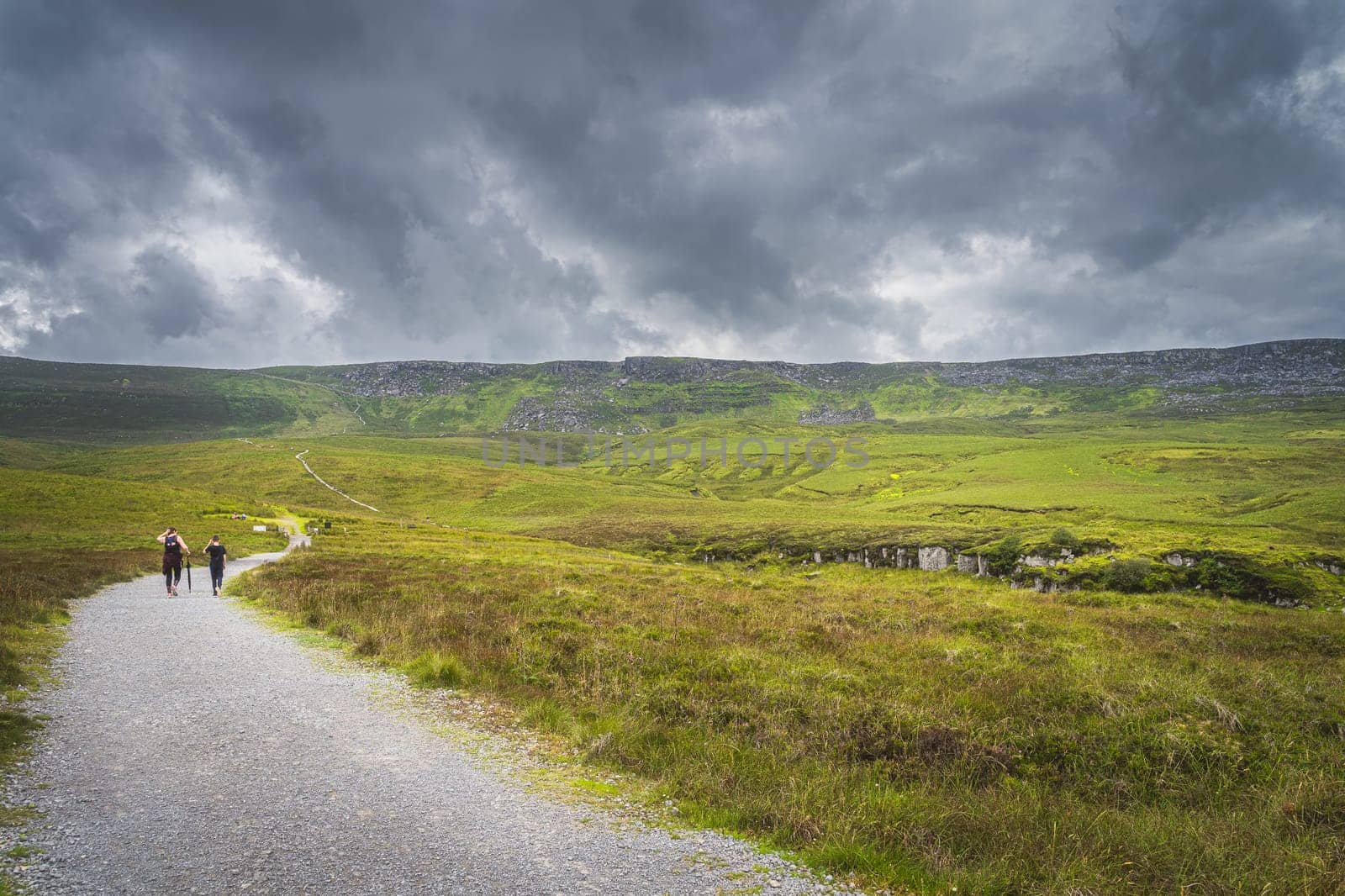 People hiking on a trail between green hills and peat bog leading to Cuilcagh Mountain with stormy, dramatic sky in background, Northern Ireland