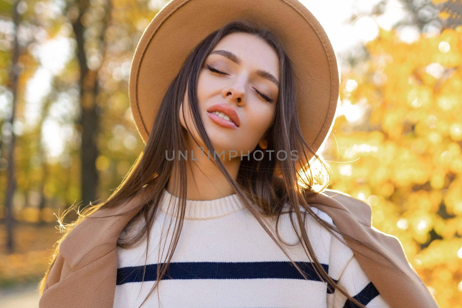 portrait of attractive stylish smiling woman with long hair walking in the park, dressed in warm brown coat autumn trendy fashion, street style wearing hat.
