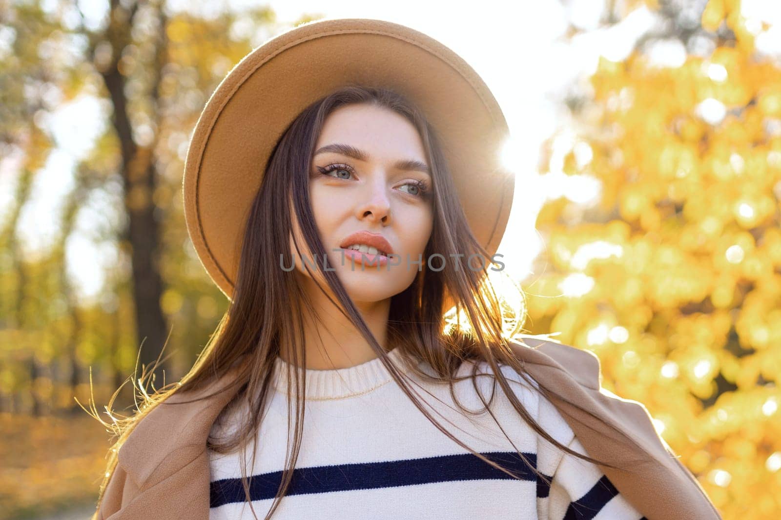 portrait of attractive stylish smiling woman with long hair walking in the park, dressed in warm brown coat autumn trendy fashion, street style wearing hat.