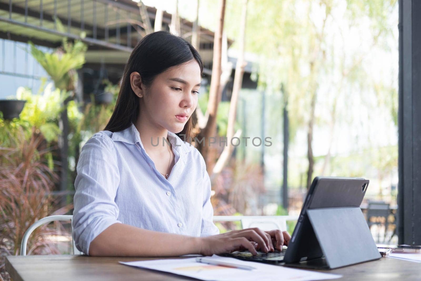 Young Asian woman freelance is working with tablet at coffee shop. Business woman typing, searching and checking document in modern cafe.  Working online technology system concept.