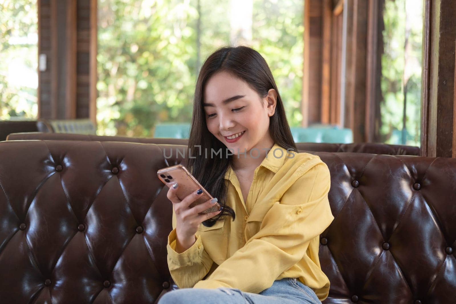 Smiling young Asian woman using smartphone at coffee shop. Asian female reading text message or social media on cell telephone during in modern cafe. Working online technology system concept.