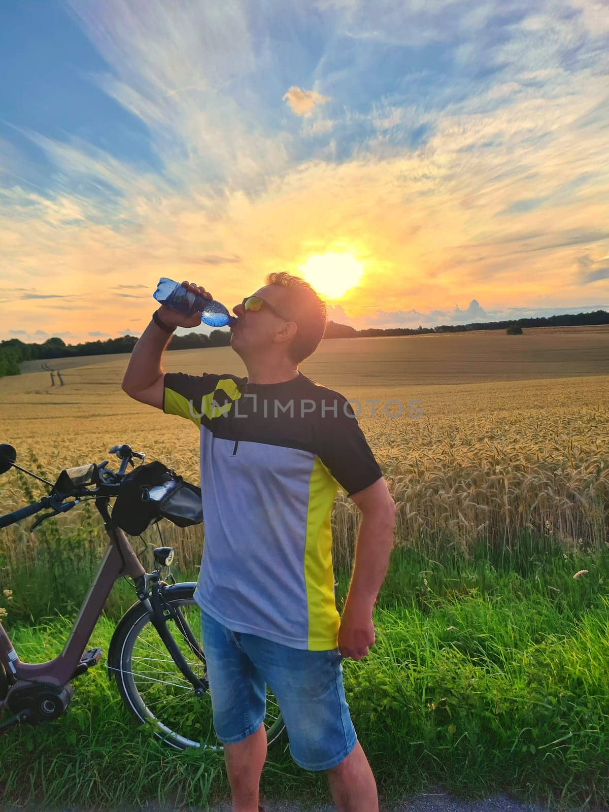 Handsome young professional cyclist dressed in cycling clothing and protective helmet feeling free and happy, drinking water from the bottle, enjoying bicycle in warm spring. Recreation of sportsman.