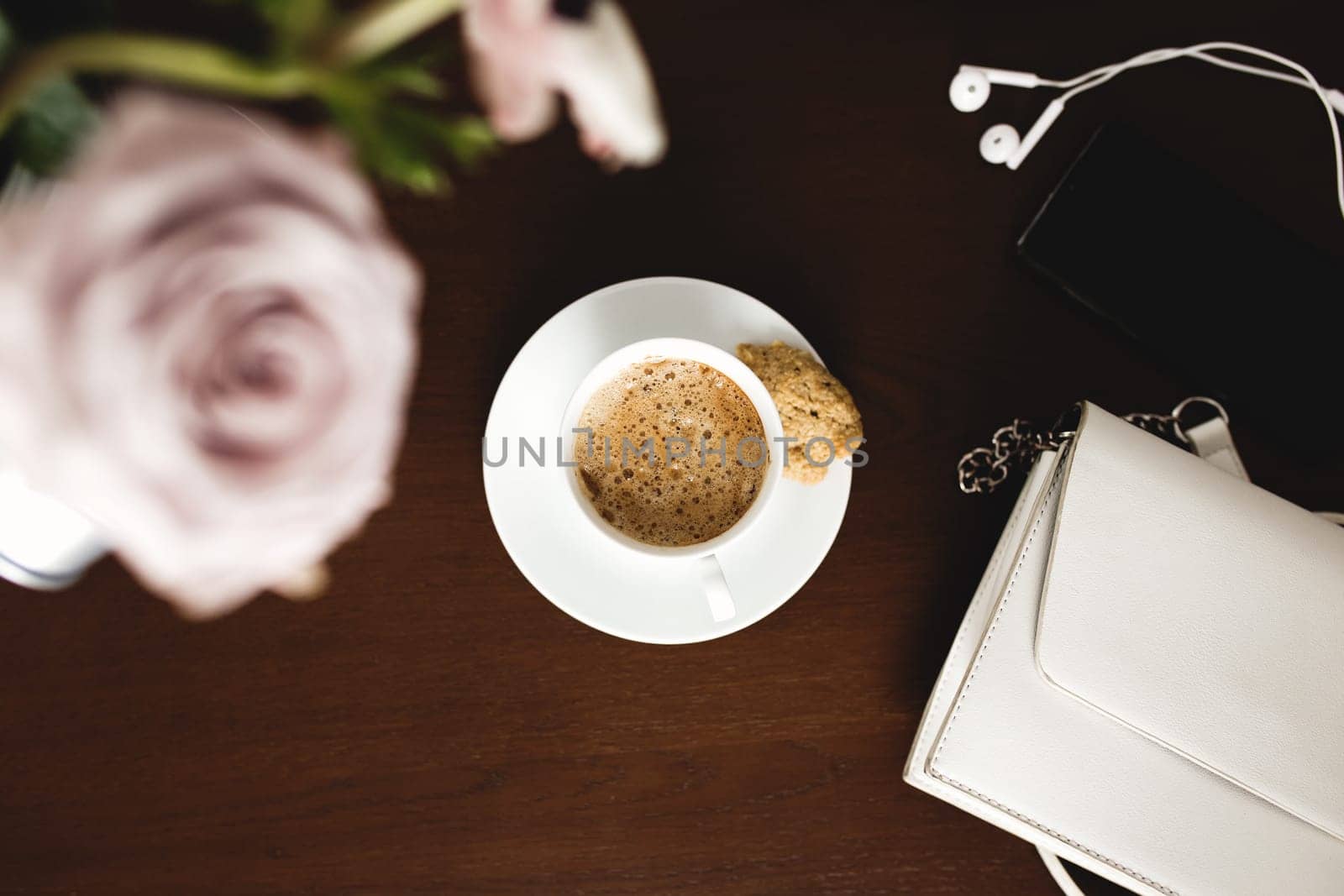 A cup of coffee with a bouquet of tender anemone, eucalyptus branches and pastel purple roses, a woman handbag, headphones and a phone on a brown wooden table. Blurred background