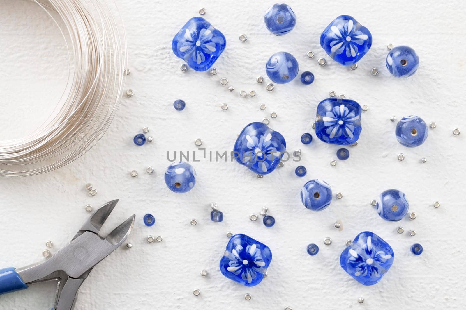 Blue and white glass beads with silver sead beads, wire cutters, and beading wire, on white painted wooden surface, top down view