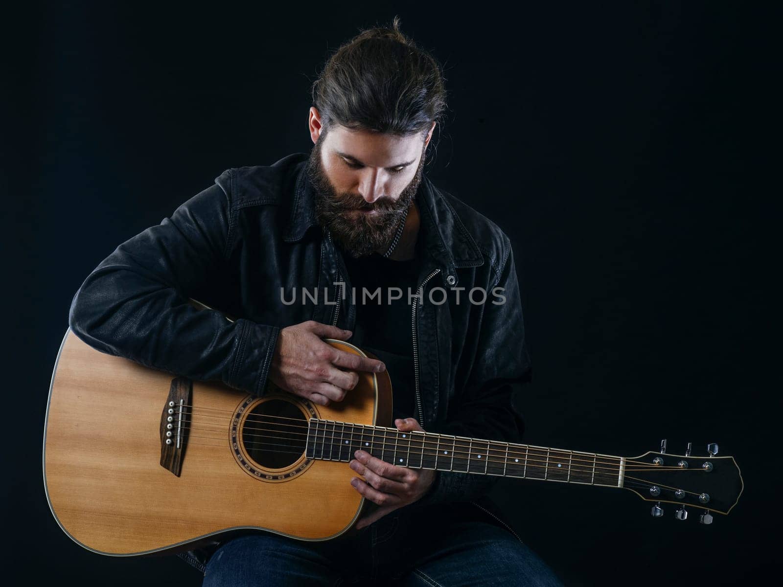Young bearded man sitting and playing an acoustic guitar over black background.