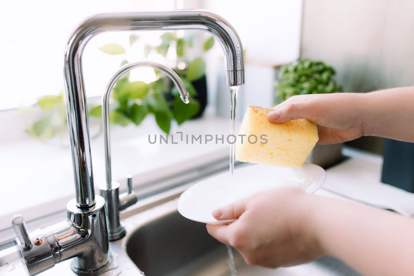 Woman hands washing white plate with sponge and soap in kitchen in sink under running water. High quality photo
