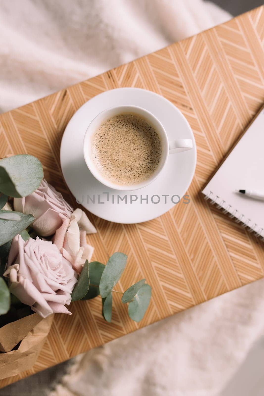 Romantic morning. Wooden coffee table with flowers on bed flat lay, coffee cup, flowers, note pad, pen. Lilac roses with eucalyptus and anemones. High quality photo