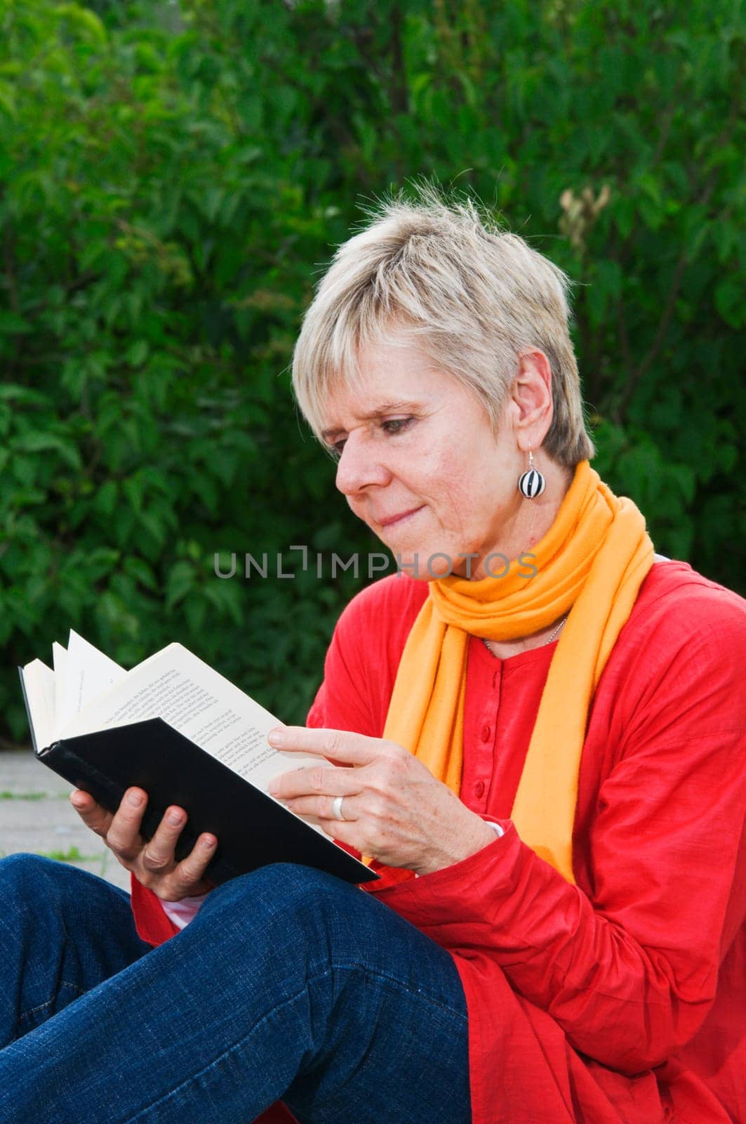 Side view of a mature woman sitting at the edge of a park and looking into an open book that is being held in front of her.