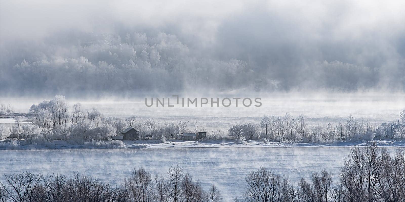 Winter frosty fog on an unfrozen river. Trees and houses in the snow on the shore. Winter landscape nature.
