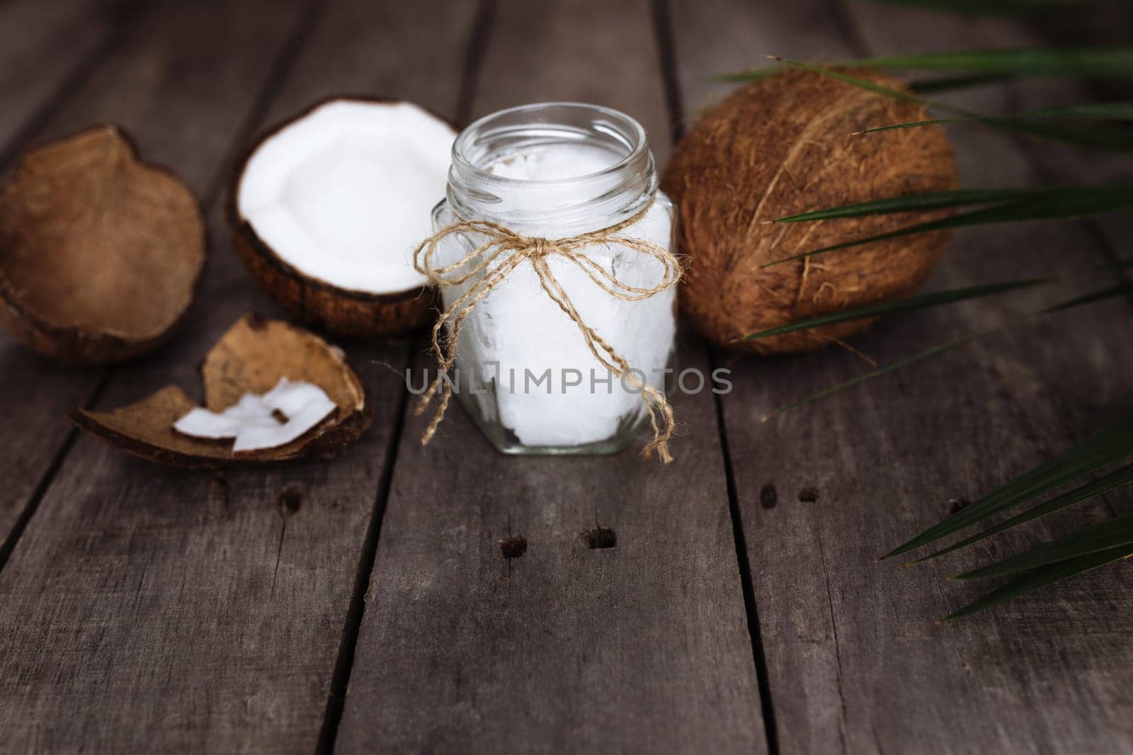 Broken coconuts on gray wooden background with jar of raw organic extra virgin coconut oil and palm leaf. White coconut pulp. High quality photo
