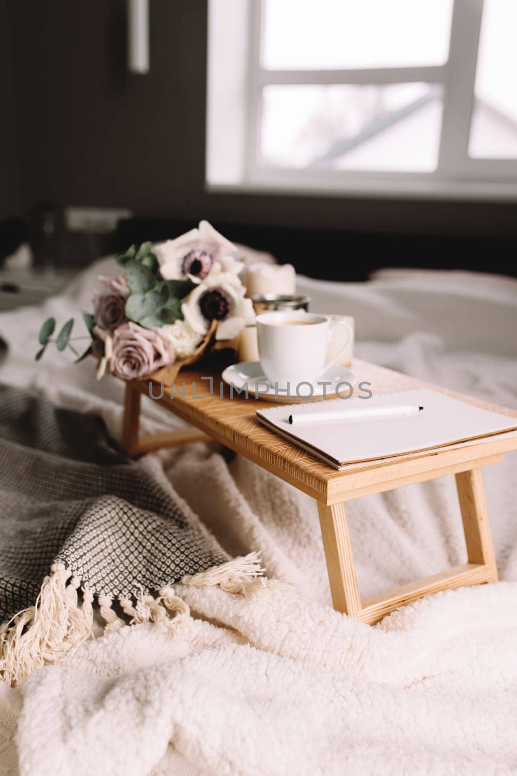 Romantic morning. Wooden coffee table with flowers on bed with plaid, coffee cup, flowers and candles, note pad, pen. Lilac roses with eucalyptus and anemones. Interior gray tones. High quality photo
