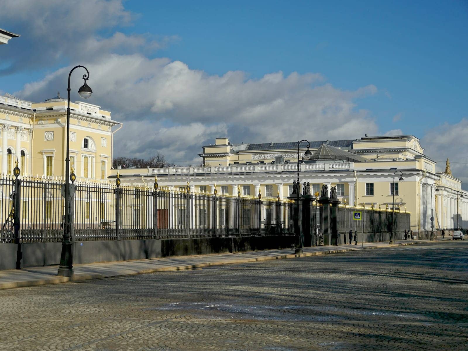 Ancient palace behind a high cast-iron fence, lonely passers-by walk along the cobbled pavement, the panorama of the old city, a sunny spring day, cumulus clouds are flying across the sky