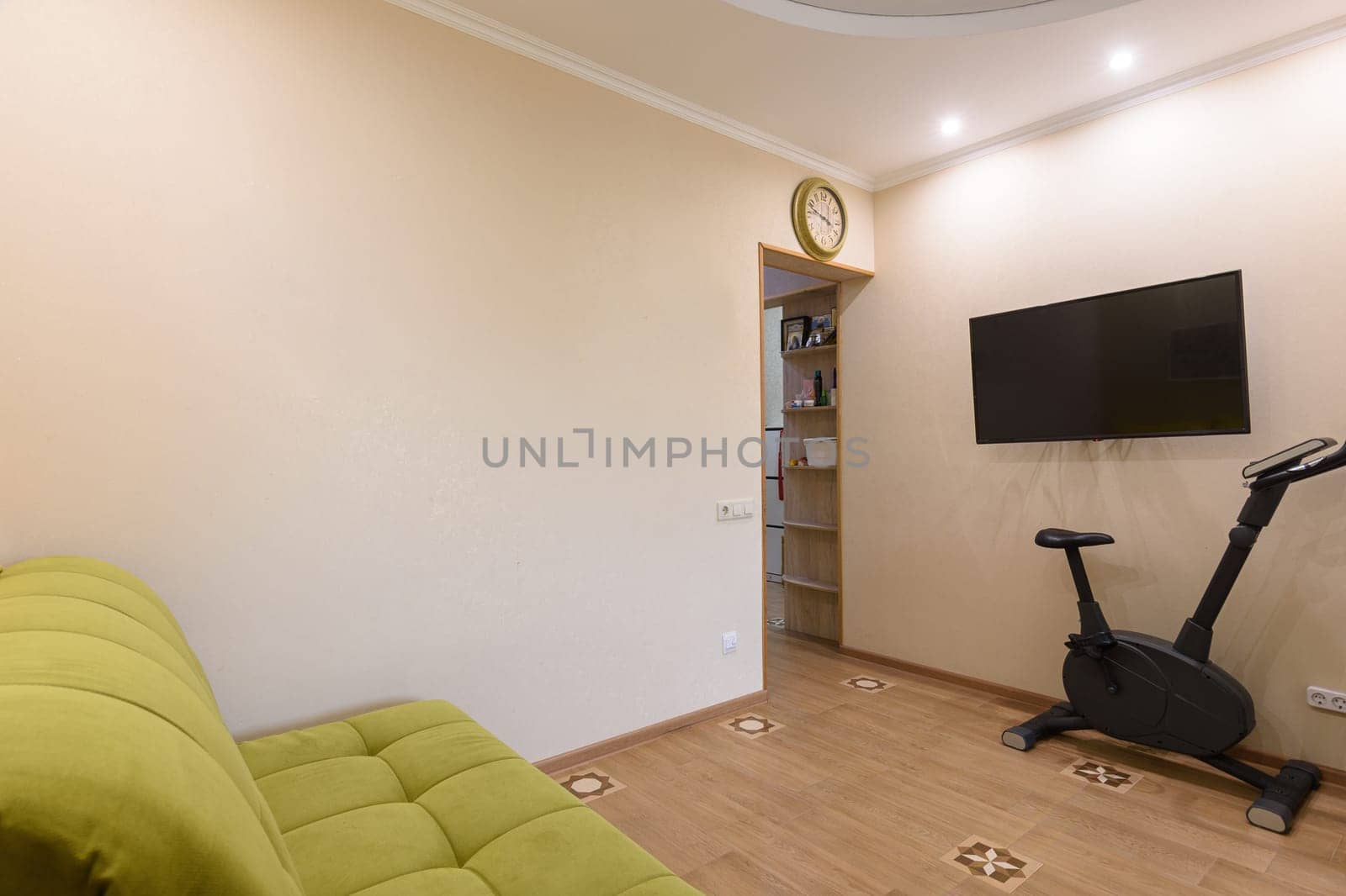 Fragment of the living room interior, in the foreground there is a sofa, on the back wall there is a TV and an exercise bike