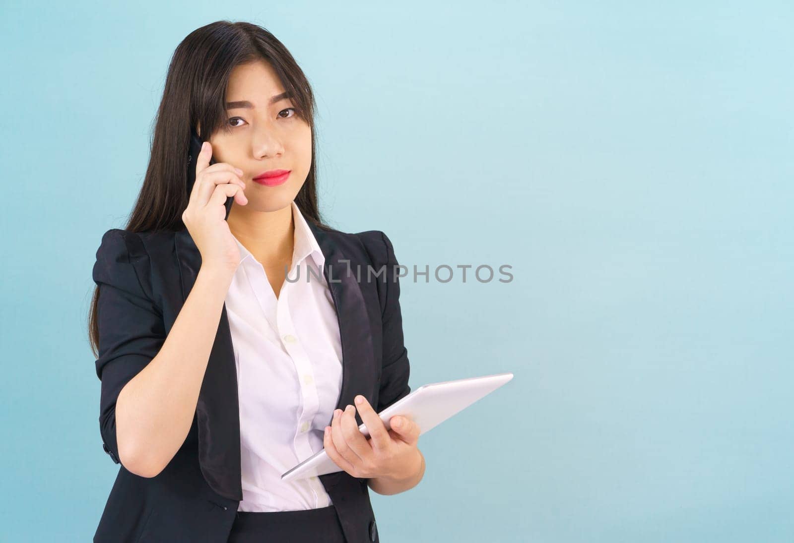 Young Asian women in suit standing using her phone and holding digital tablet against blue background