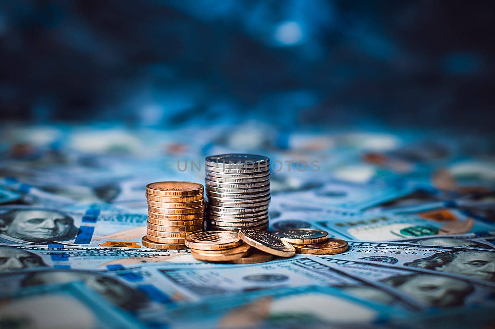 Stacks of coins against the background of hundred-dollar bills are scattered throughout the space. The coins are collected in two columns, they are shiny, golden in color.