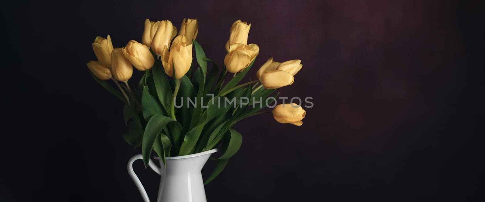 Tulips in a jug. Classic still life with a bouquet of yellow tulip flowers in a vintage white jug on a dark background and an old wooden table. Art photography.