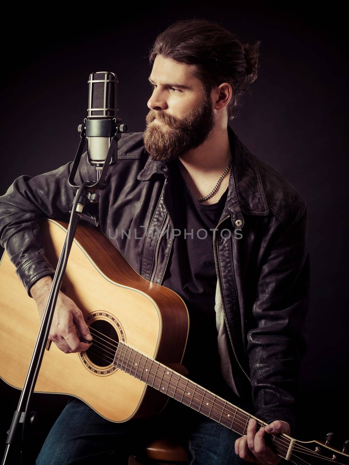 Young bearded man sitting and playing an acoustic guitar over black background..