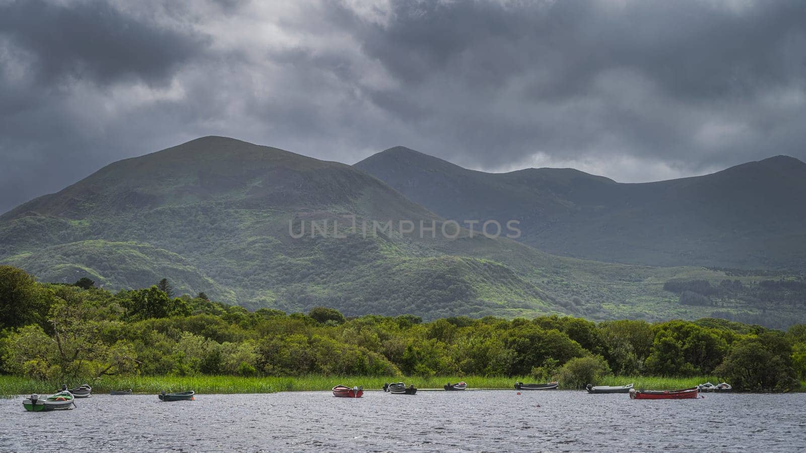 Wooden boats moored on Lough Leane with mountains of Killarney National Park and dark dramatic clouds in background, Ring of Kerry, Ireland