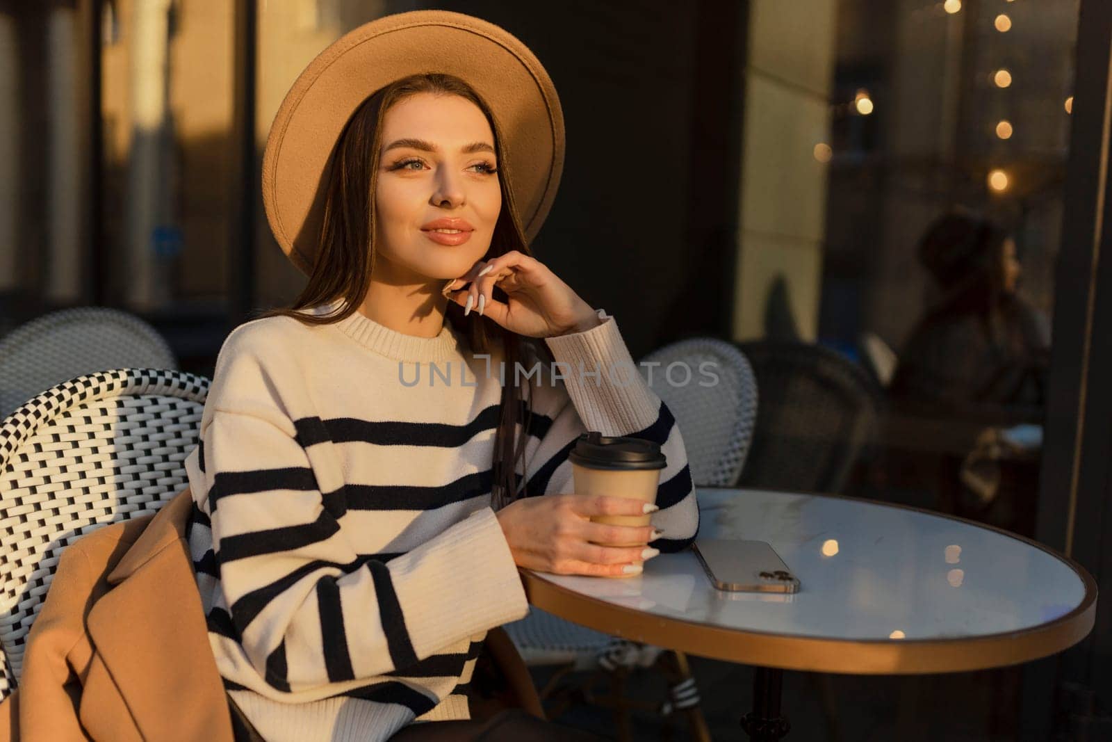 beautiful smiling woman in stylish outfit sitting at the table in a hat and sweater, romantic happy mood, waiting for a boyfriend on a date in a cafe, spring-summer fashion trend, drinking coffee.