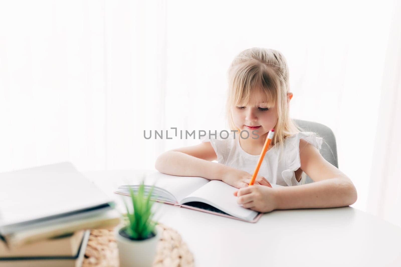 A little girl sits at the table and writes in a white notebook. Education concept. Home schooling. Homework. High quality photo