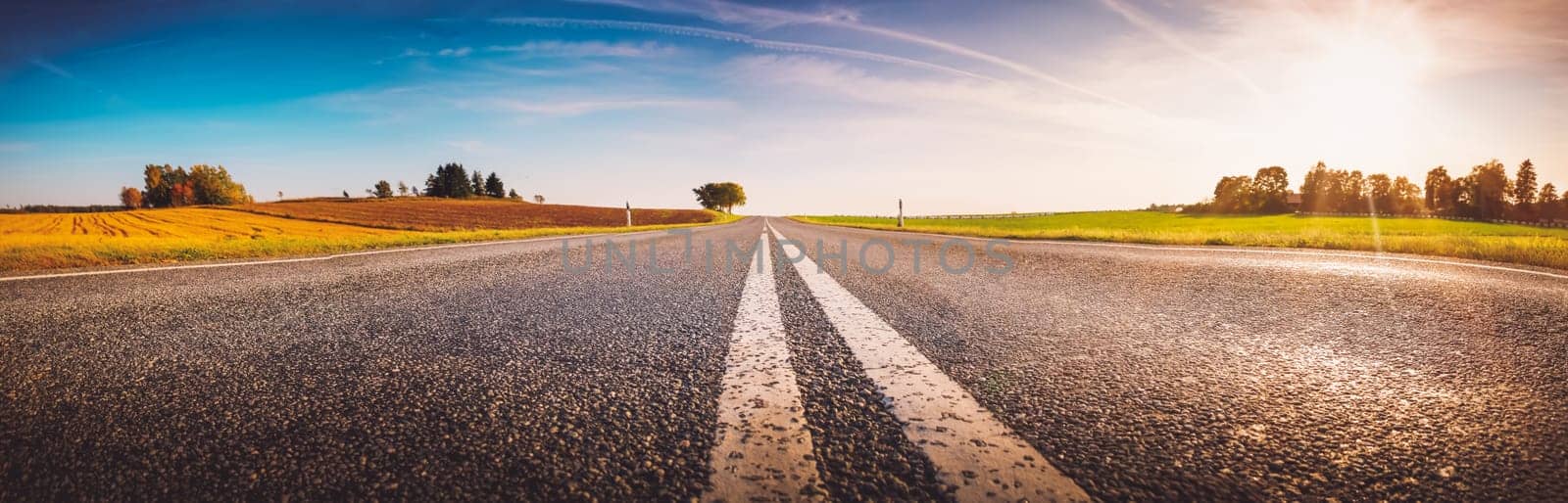 Wide asphalt road panorama in countryside in sunset on summer. Route with white dividing lines in beautiful nature landscape.