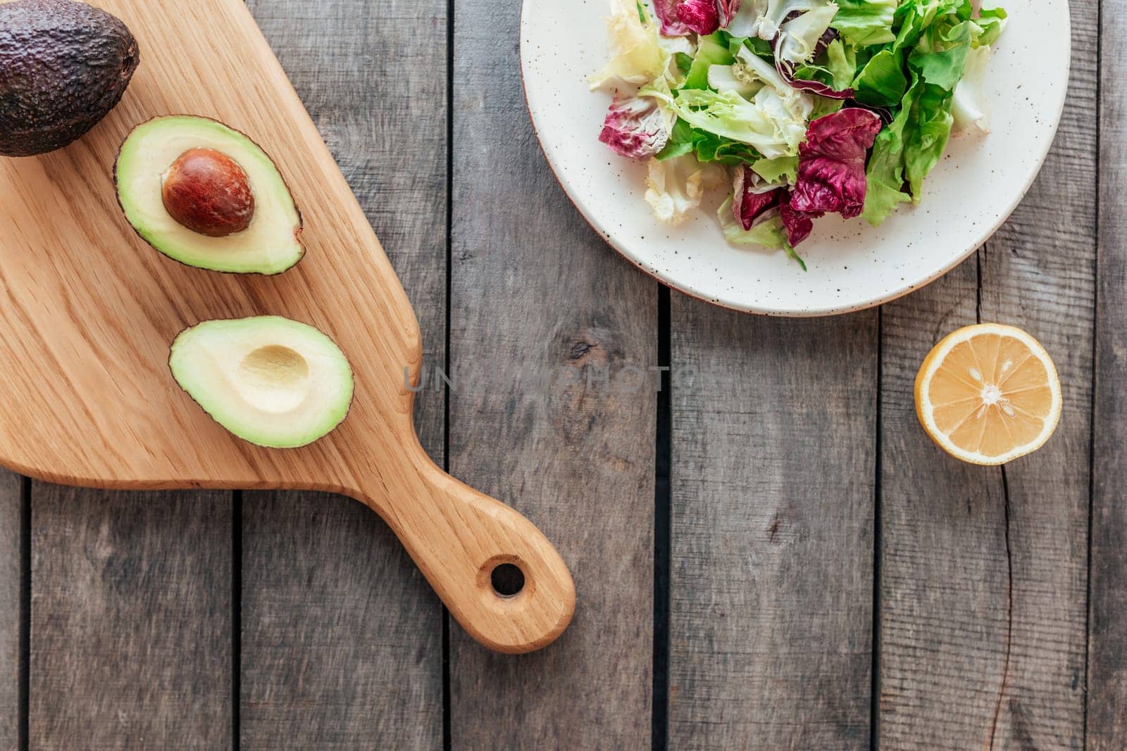 Healthy eating concept flat lay. Mediterranean diet, plate with fresh green lettuce salad leaves, purple radicchio leaves, cutting board with avocado halves, kernel, lemon on wooden background