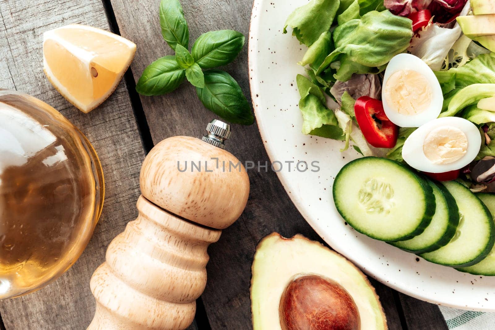 Healthy eating concept flat lay. Mediterranean diet, plate with lettuce salad leaves, quail eggs, avocado half with kernel, cucumber slices, pepper, basil, olive oil, lemon, salt on wooden background