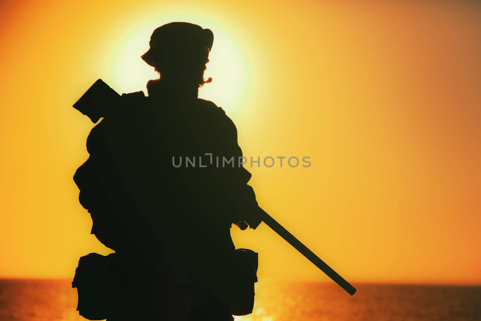 Silhouette of army elite forces sniper, commando shooter in boonie hat, carrying backpack, equipped tactical radio, walking on background of setting or raising sun. Counter terrorist forces soldier