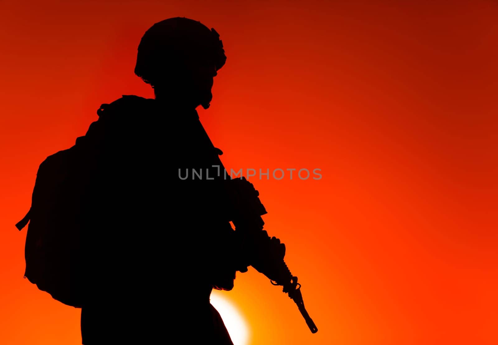 Silhouette of army rifleman in helmet and ammunition, carrying tactical backpack, walking with service rifle on background of sunset sky. Counter terrorist forces fighter marching at night mission