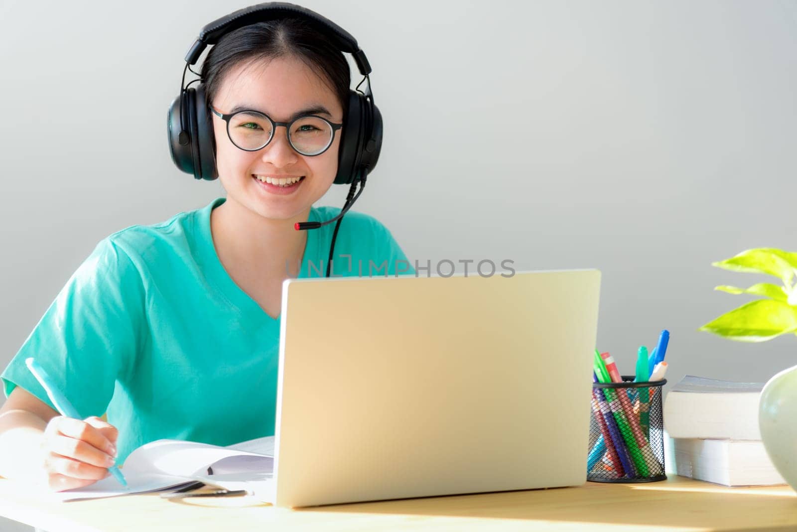 Portrait young Asian female student with headphones glasses smile looking up a note on the book a teenage girl is happy to university internet distance study online class on a laptop computer at home