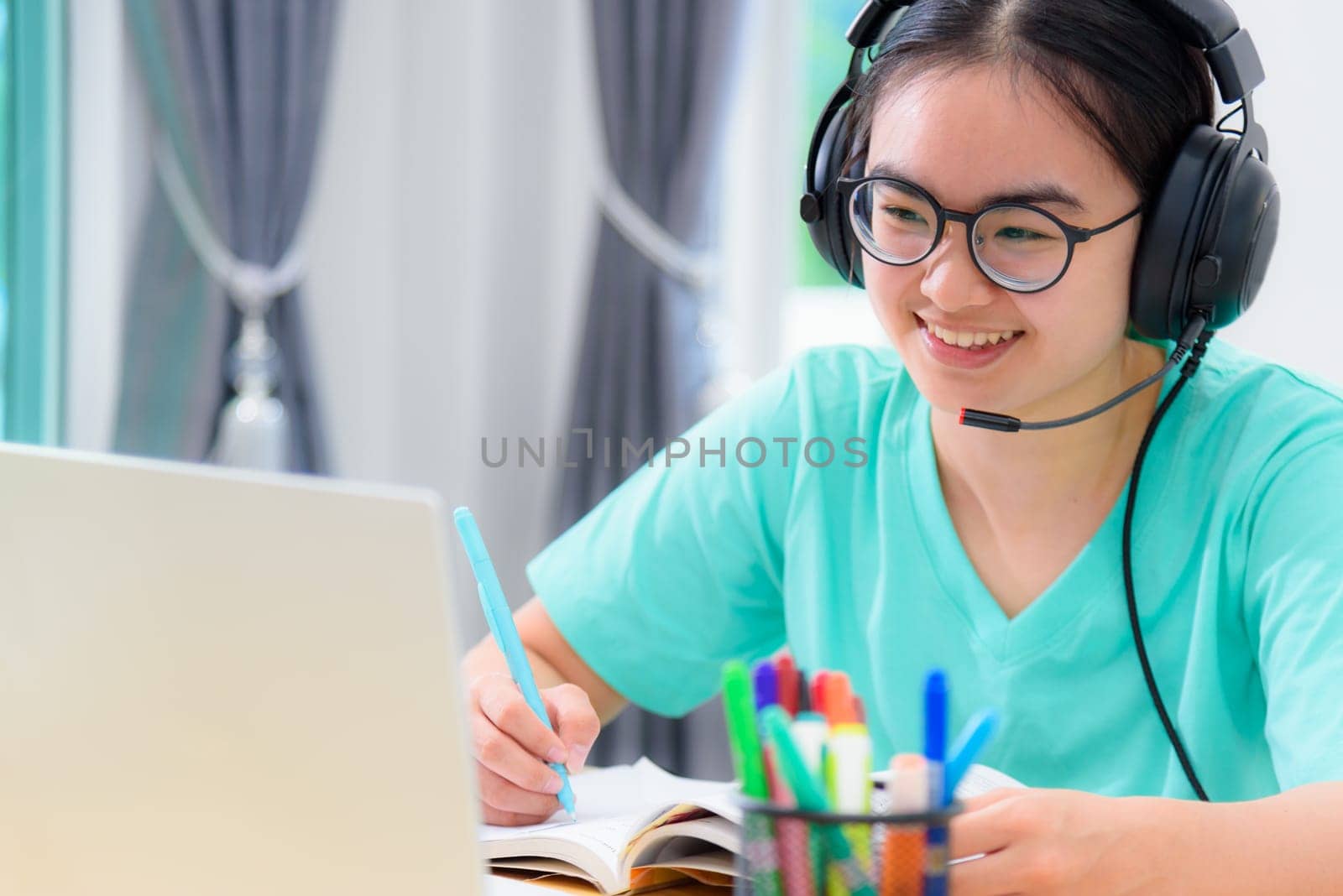 Young Asian woman student with headphones and glasses writing on the book looking at video meeting on laptop computers a girl is happy to take the university internet distance learning online class