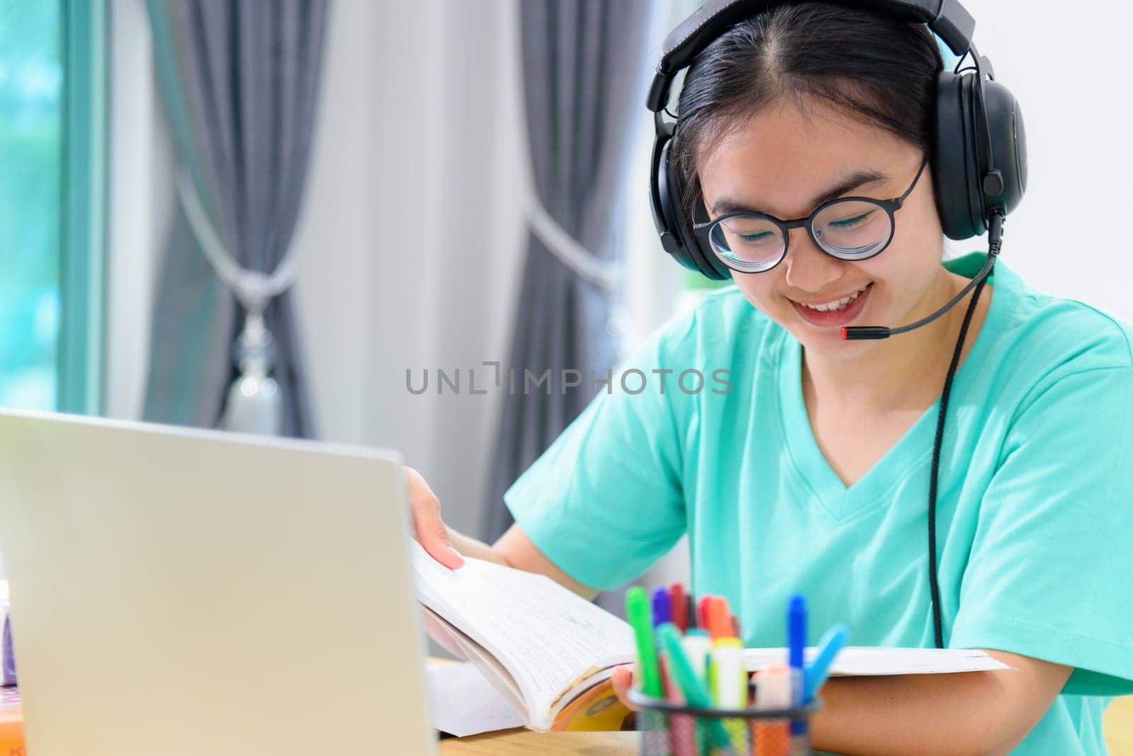 Portrait young Asian woman student with headphones glasses is smiling reading a book study a teenage girl is happy to college internet distance learning online class works on a laptop computer at home