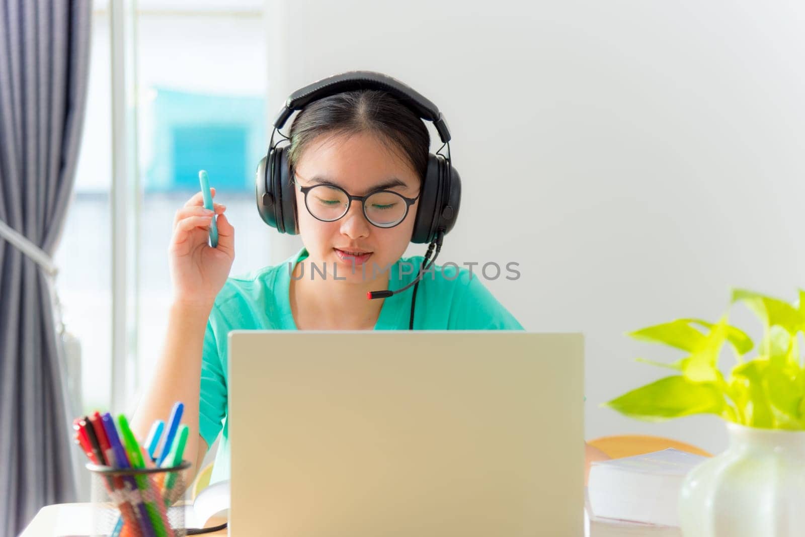 Asian young woman student with glasses headphones girl study serious writing note on book looking video conference laptop computer university class online internet learning distance education at home