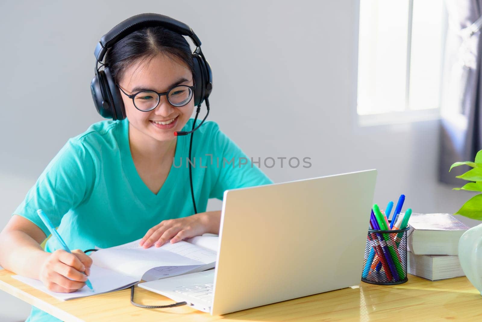 Asian young woman student with glasses headphones girl study happy writing note on a book looking video conference laptop computer university class online internet learning distance education at home