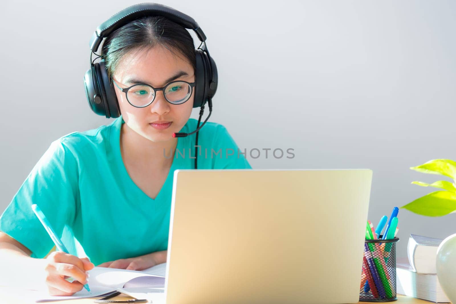 Asian young woman student with glasses headphones study serious writing note on a book looking video conference on laptop computer university class online internet learning distance education at home