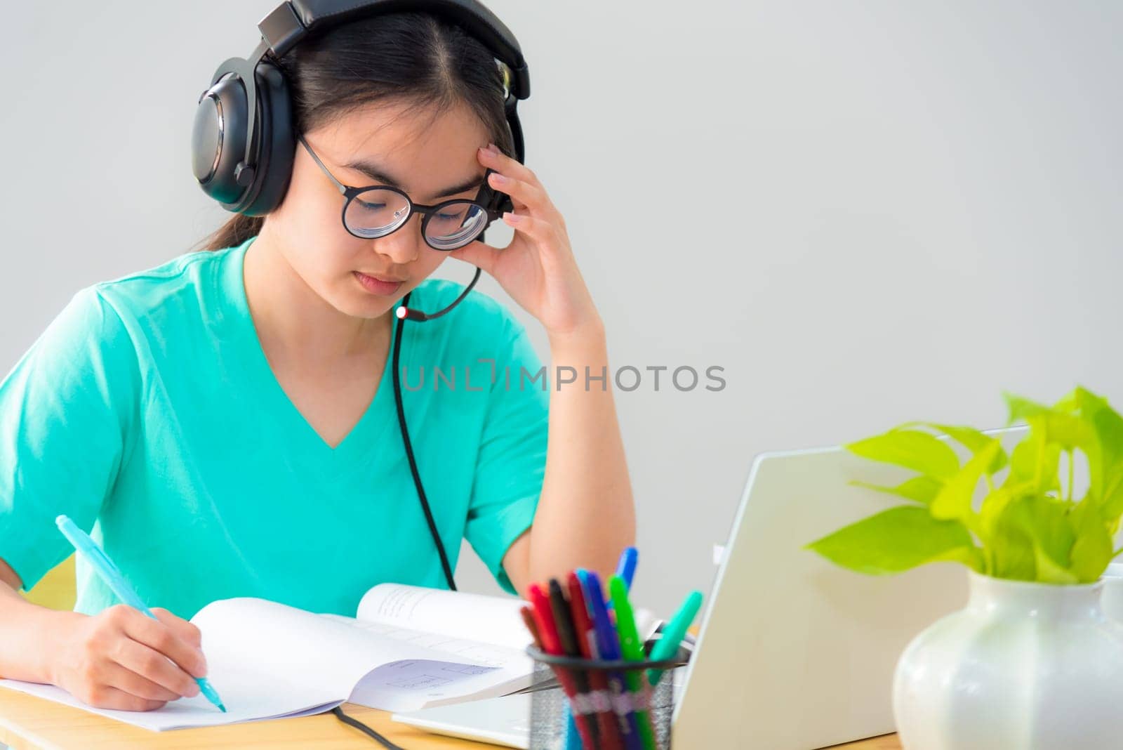 Asian woman student teenage girl with glasses headphones sitting looking serious reading a book worry using laptop computer on table learning online study. Education from a class of university at home