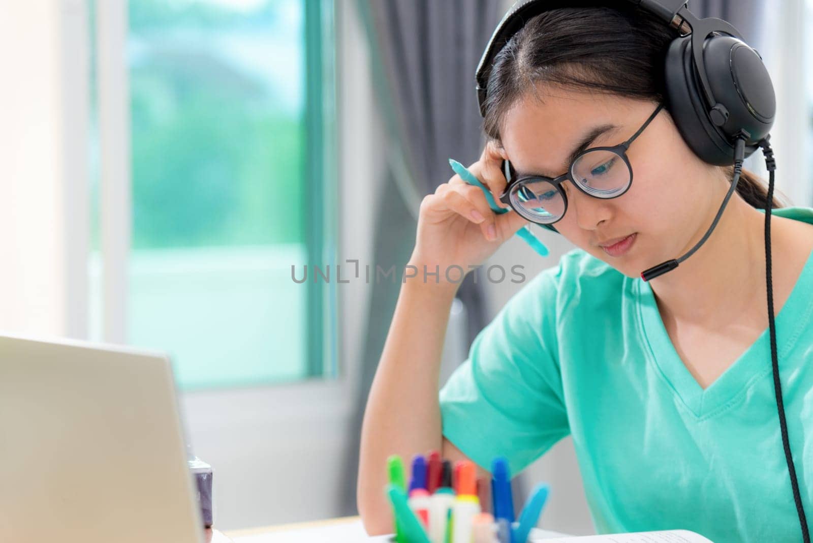 Asian woman student teenage girl with glasses headphones sitting looking serious reading a book worry using laptop computer on table learning online study. Education from a class of university at home